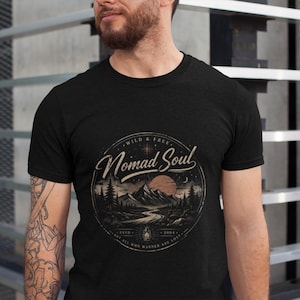 May include: Black t-shirt with a circular graphic featuring a mountain landscape, river, and sunset. The text reads "Wild & Free Nomad Soul" with additional text and a small lantern illustration. The shirt is short-sleeved.