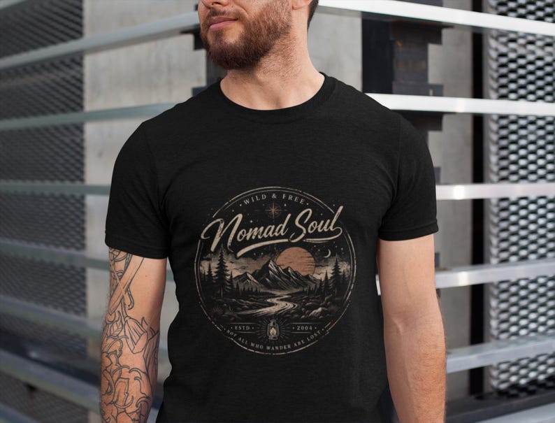 May include: Black t-shirt with a circular graphic featuring a mountain landscape, river, and sunset. The text reads "Wild & Free Nomad Soul" with additional text and a small lantern illustration. The shirt is short-sleeved.