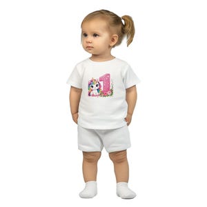 Peut inclure: Ensemble blanc pour tout-petit composé d'un t-shirt et d'un short. Le t-shirt présente un motif de licorne colorée avec le chiffre "1" et des détails floraux, parfait pour une fête de premier anniversaire.