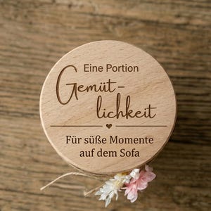 Könnte beinhalten: Ein runder, heller Holzdeckel mit der Gravur "Eine Portion Gemütlichkeit" über einem kleinen Herz und dem Text "Für süße Momente auf dem Sofa," verziert mit kleinen weißen und rosafarbenen künstlichen Blüten am unteren Rand.