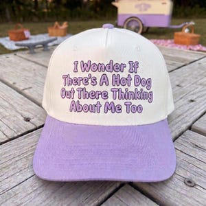 May include: A baseball cap with a white crown and a light purple brim. The front of the crown features purple embroidered text that reads, I Wonder If There's A Hot Dog Out There Thinking About Me Too, in a rounded, playful font style.