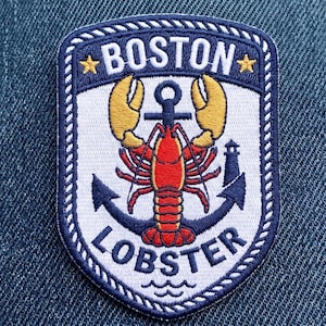 May include: Embroidered patch featuring a red lobster with yellow claws, an anchor, and a lighthouse. The patch is white with a navy blue border and the words "BOSTON LOBSTER" in white lettering. Two gold stars are at the top.