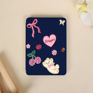 Può includere: Una custodia rettangolare per e-reader di colore blu scuro con una trama diagonale simile al denim. La custodia presenta vari adesivi, tra cui un fiocco rosa, una farfalla, un cuore con la scritta Babygirl, labbra rosse, una fragola, ciliegie e un orsetto color crema.