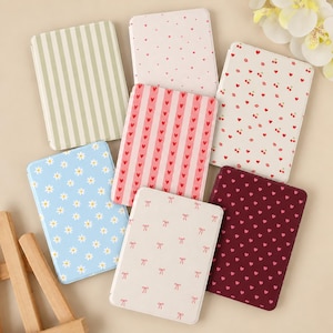 May include: A collection of seven rectangular compact mirrors featuring various patterns including sage green stripes, pink hearts, red cherries, blue daisies, and white bows on a cream background, arranged in a scattered layout.