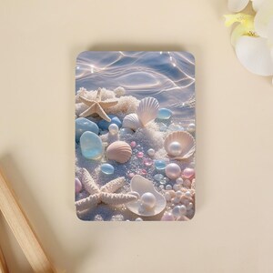 Pode incluir: Uma impressão retangular com uma cena de praia com areia branca, duas estrelas-do-mar bege, várias conchas brancas e rosas, seixos de vidro azul e rosa espalhados, juntamente com várias pérolas esféricas brancas.