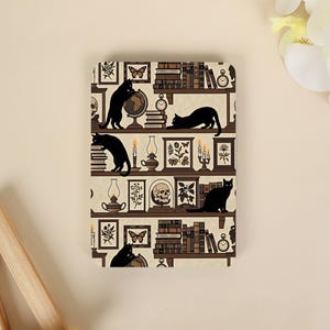 May include: A rectangular notebook cover featuring a repeating pattern of black cats, stacks of books, globes, skulls, framed botanical illustrations, and lit candles on shelves against a cream-coloured background with a subtle wallpaper texture.