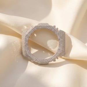 May include: A circular watch bezel accessory featuring a silver-toned metal frame completely encrusted with numerous small, clear, sparkling stones. The piece has a distinct octagonal outer shape with protruding edges and a smooth, circular inner opening.