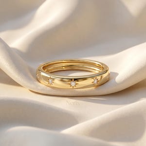 May include: A gold band ring featuring multiple star-shaped engravings, each centred with a small, clear, sparkling stone. The ring has a polished, reflective surface and is positioned on a soft, draped, off-white fabric. The text Nerix is visible.