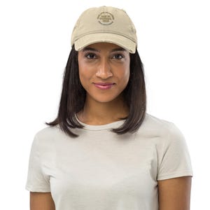May include: A beige distressed baseball cap featuring the text "The Best Is Yet To Come" in a circular design. The cap is worn by a person wearing a white t-shirt.