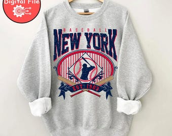 May include: A light gray sweatshirt with the words "Baseball New York" in navy blue and red. The design includes a baseball player silhouette, crossed bats, and the text "EST 1903". The sweatshirt is hanging on a wooden hanger.