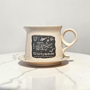 May include: A beige ceramic mug with a matching saucer. The mug features a black rectangular design with white illustrations of a sun, fish, and other abstract shapes. The handle is a loop shape.