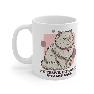 May include: A white ceramic mug featuring a graphic illustration of a fluffy grey cat wearing a pink jewelled collar. The cat has a grumpy expression. Below the cat, the text reads: EXPENSIVE, DIFFICULT, & TALKS BACK in dark purple lettering.