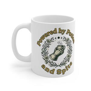 May include: A white ceramic mug featuring a graphic of a metal gauntlet holding a steaming cup, surrounded by a floral wreath and moon phases. The text "Powered by Potion and Spite" is printed in gold lettering around the central illustration.