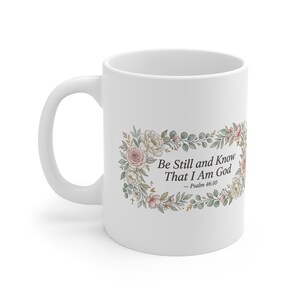 Pode incluir: Caneca de cerâmica branca com o texto "Be Still and Know That I Am God" dentro de uma grinalda floral. A grinalda inclui flores rosa e creme com folhas verdes. A caneca tem uma alça curva.