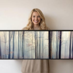 May include: A framed landscape art print featuring a misty forest scene with tall, bare trees in shades of blue, gray, and beige. The artwork is displayed in a black frame, creating a serene and natural aesthetic.
