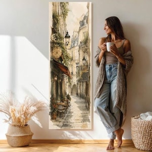 Könnte beinhalten: Ein hohes, vertikales, rechteckiges Wandkunstbild, das eine schmale europäische Straßenszene mit Steingebäuden, einem Café mit Sitzgelegenheiten im Freien, hängenden Laternen und viel Grün zeigt. Das Kunstwerk besticht durch eine gedeckte Farbpalette in Beige, Braun und Grün.