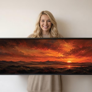 Op de afbeelding: Een panoramisch landschapsschilderij in een dunne zwarte lijst met een levendige zonsondergang boven een verre bergketen en een kalm wateroppervlak. De lucht is gevuld met dramatische wolken in tinten van diep oranje, vurig rood en donker karmijn.