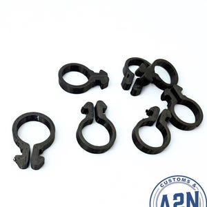 以下が含まれることがあります： 白い表面に散らばる、黒い3Dプリントされたプラスチッククリップがいくつかあります。 クリップは、留め金が付いた円形デザインです。 画像には、「CUSTOMS & A2N 3D STUDIO」というテキストが入った円形のロゴも含まれています。