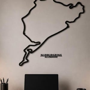 May include: A black metal wall art piece depicting the Nürburgring race track layout. The design features a continuous outline of the circuit with the text NURBURGRING ALLEMAGNE positioned below the track shape in a bold, sans-serif font.