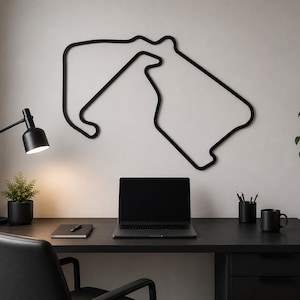 May include: A black metal wall art piece depicting a race track circuit hangs above a desk. The desk is black and has a laptop, a pen holder, a mug, and a small plant. A black desk lamp and a chair complete the workspace.