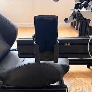 Peut inclure: Support de téléphone noir fixé à un siège de simulateur de course noir. Le support de téléphone est monté sur un cadre métallique noir. Le siège est noir avec un motif texturé. L'image est prise à l'intérieur.