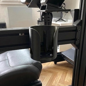 May include: Black cup holder attached to a black racing simulator rig. The rig includes a steering wheel, seat, and monitor mount. The cup holder is mounted on the side of the rig, near the seat. The background shows a room with a window and wooden floor.