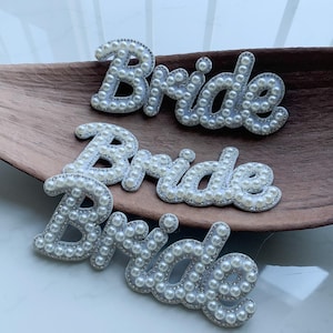 Pode incluir: Três recortes decorativos com a palavra "Bride". Cada palavra é escrita com pérolas de imitação brancas, com uma borda prateada. Os recortes são dispostos em uma bandeja de madeira marrom em forma de folha.