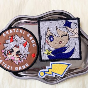 May include: Embroidered patches featuring anime-style characters and designs. One patch is round with the text "ARATAKI GANG" and a character illustration. Another is square with a girl winking. A yellow lightning bolt patch is also included.