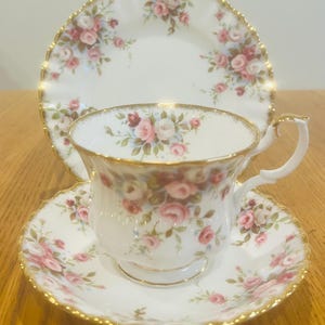 May include: A white teacup, saucer, and plate set with a floral rose pattern in pink and red. The edges are trimmed with gold. The teacup has a curved handle and sits on a matching saucer. A larger plate is behind the teacup.