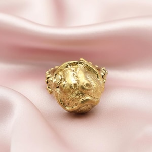 May include: A gold-colored ring with a textured, sculpted design of a face. The ring is set against a soft, pink background, highlighting its intricate details and metallic sheen. The ring's design features a detailed face with a slightly sad expression.