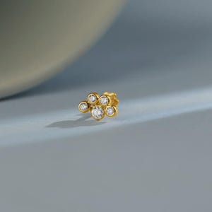 Könnte beinhalten: Goldener Ohrstecker mit sechs klaren Edelsteinen. Der Ohrring hat ein Cluster-Design, mit einem größeren zentralen Stein, der von kleineren Steinen umgeben ist. Der Hintergrund ist ein weiches, gedämpftes Blau. Das Design ähnelt einem Pfotenabdruck oder einer Blume.