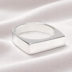 May include: A polished silver ring with a square face and a rounded inner band. The ring is displayed on a white, textured fabric. The ring's design is minimalist and contemporary.