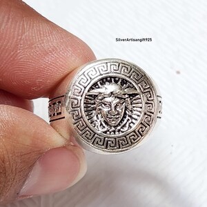 May include: A silver ring with a detailed Medusa head design within a circular frame. The ring has a textured surface and a band with geometric patterns. The ring is held up by a hand.