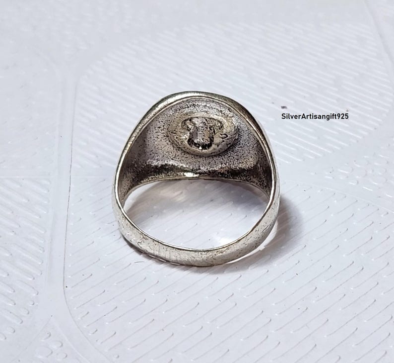 May include: A silver ring featuring a detailed emblem on a circular face. The ring has a thick band and a slightly textured surface. The emblem appears to be a detailed design. The ring is set against a white background.