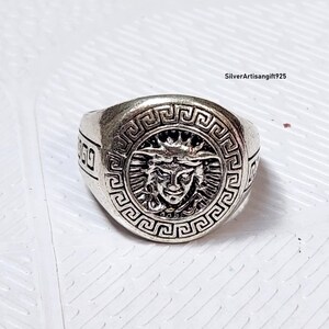 May include: A silver ring with a Medusa head design at the centre. The ring has a Greek key pattern around the Medusa image and on the band. The ring is likely made of metal.