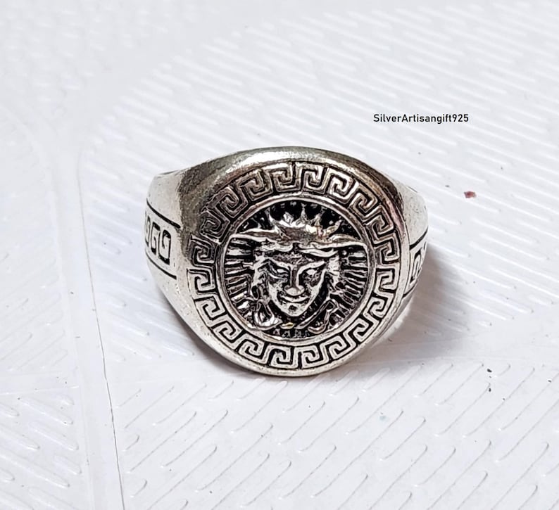 May include: A silver ring with a Medusa head design at the centre. The ring has a Greek key pattern around the Medusa image and on the band. The ring is likely made of metal.