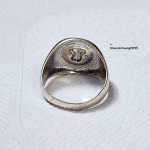 May include: A silver ring featuring a detailed emblem on a circular face. The ring has a thick band and a slightly textured surface. The emblem appears to be a detailed design. The ring is set against a white background.