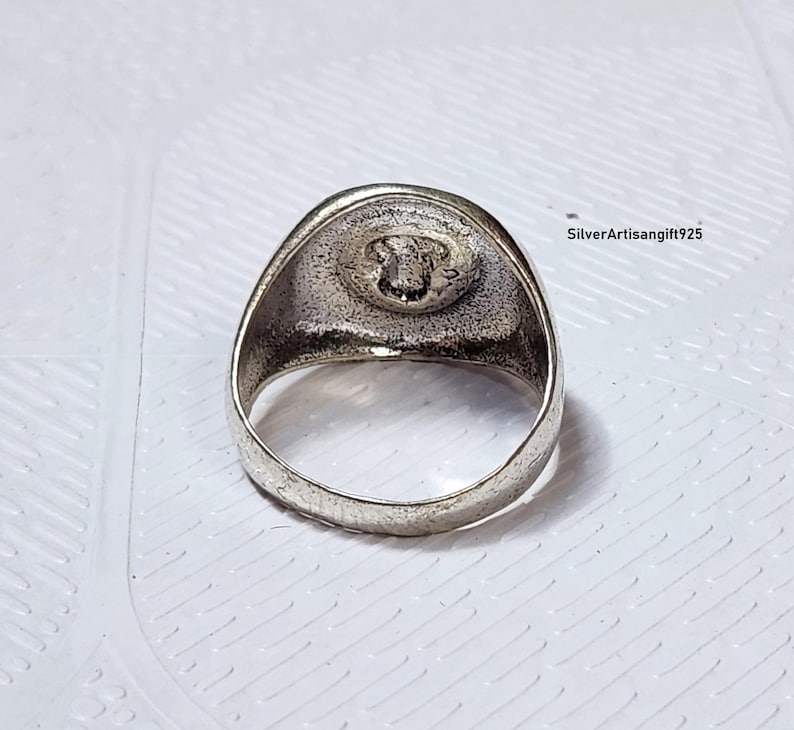May include: A silver ring featuring a detailed emblem on a circular face. The ring has a thick band and a slightly textured surface. The emblem appears to be a detailed design. The ring is set against a white background.