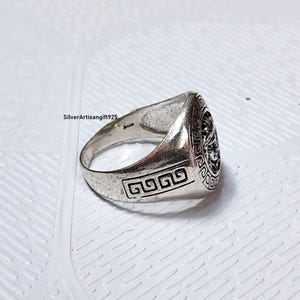 May include: A silver ring featuring a detailed face design on the top. The ring has a rectangular design on the side. The ring is on a white surface. The text "SilverArtisanGift925" is visible.