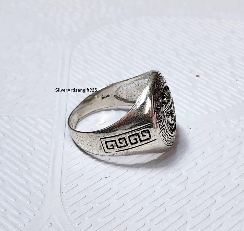 May include: A silver ring featuring a detailed face design on the top. The ring has a rectangular design on the side. The ring is on a white surface. The text "SilverArtisanGift925" is visible.