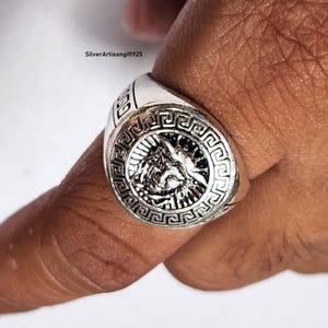 May include: A silver ring with an intricate design. The ring has a central emblem with a figure and sunburst pattern, framed by a Greek key border. The ring is on a finger.