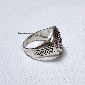 May include: A silver ring featuring a detailed face design on the top. The ring has a geometric pattern on the side, set against a white textured surface. This is a piece of jewellery.