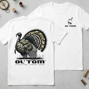 Könnte beinhalten: Zwei weiße T-Shirts mit Truthahn-Motiven. Eines zeigt eine detaillierte Illustration eines Truthahns mit einem Tarnmuster-Schwanz und dem Text "OL' TOM TECHNICAL TURKEY GEAR". Das andere hat ein kleineres Logo mit zwei Truthähnen und dem Text "OL' TOM".