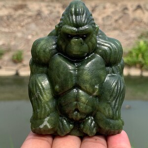 May include: A detailed, green jade gorilla sculpture. The gorilla is depicted in a seated position, with a muscular physique and a stern expression. The sculpture is carved with intricate details, showcasing the texture and color variations of the jade.