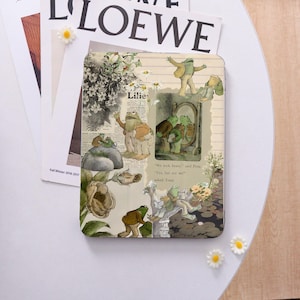 Könnte beinhalten: Eine dekorative Metallbox mit Illustrationen von Fröschen in verschiedenen Szenen, mit dem Text "Lilies" und Zitaten aus einer Geschichte. Die Box ist mit floralen und botanischen Elementen verziert und vor dem Hintergrund eines Magazins platziert.