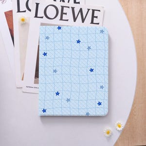 May include: A light blue notebook with a grid pattern and scattered blue star shapes. The notebook is resting on a white surface with a magazine and small decorative flowers.