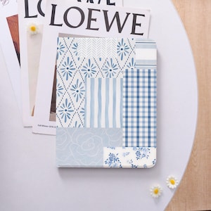 May include: A blue and white patterned notebook with various designs, including floral, striped, and checkered patterns. The notebook is rectangular and lies on top of a magazine. Small white daisy decorations are scattered around the notebook.