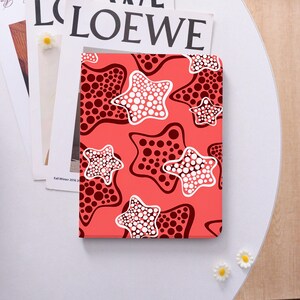 May include: A rectangular tablet case featuring a vibrant coral-red background patterned with stylized star shapes. Each star is filled with a dense arrangement of white or dark maroon circular dots, creating a repeating graphic design across the surface.