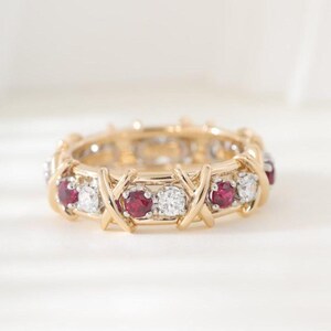 May include: A yellow gold band ring featuring an alternating pattern of round-cut red rubies and clear white diamonds. The metal setting incorporates a repeating X-shaped design that secures each gemstone within the circular structure of the ring.