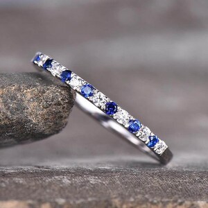 May include: A thin silver-toned band ring featuring an alternating pattern of small, round, brilliant-cut clear stones and deep blue sapphires set along the top half of the metal band, creating a continuous line of sparkling gemstones.